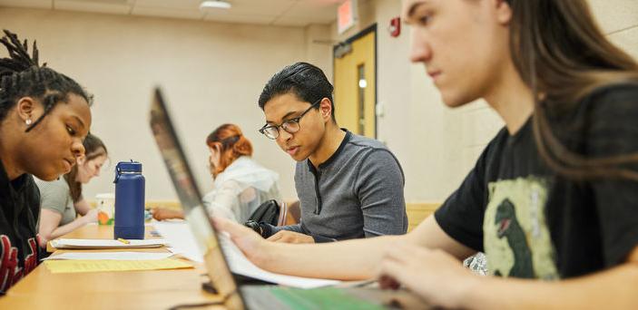 Students sit at a long desk working in groups 